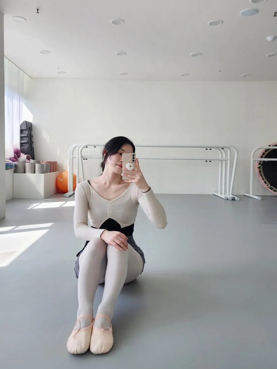 A woman in a cream ballet wrap top, tights and pink canvas ballet shoes sitting on a grey studio floor by the barres