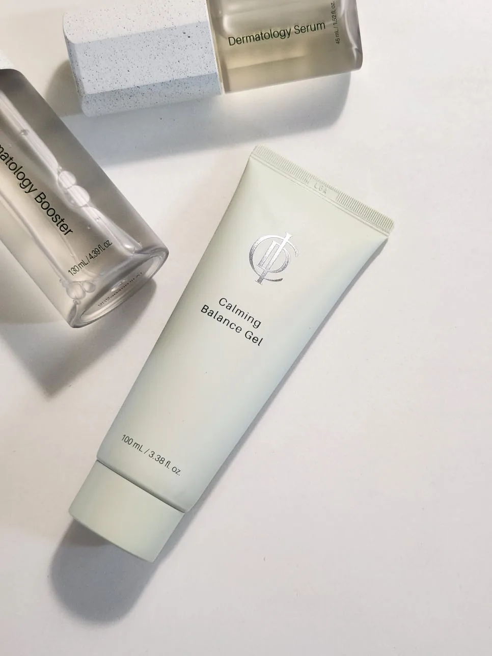 The sage-green Calming Balance Gel tube lying on white beside clear ICD Dermatology Booster and Serum bottles