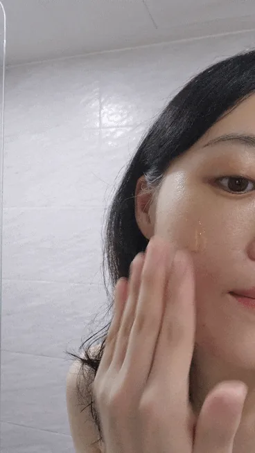 A woman spreading clear gel across her cheek with her fingertips in front of a white-tiled bathroom wall