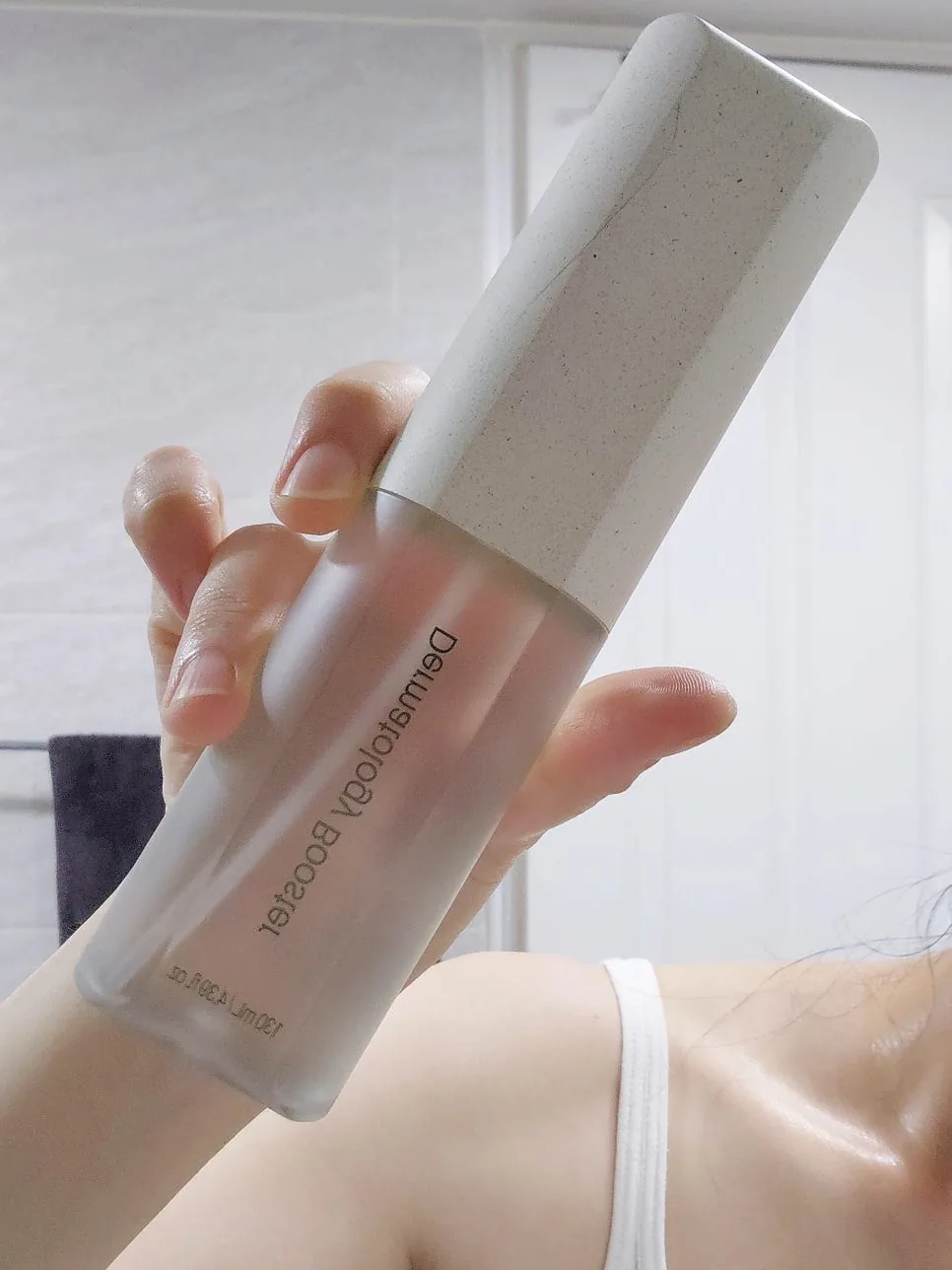Hand holding the frosted Dermatology Booster bottle with its speckled cap in a bright bathroom