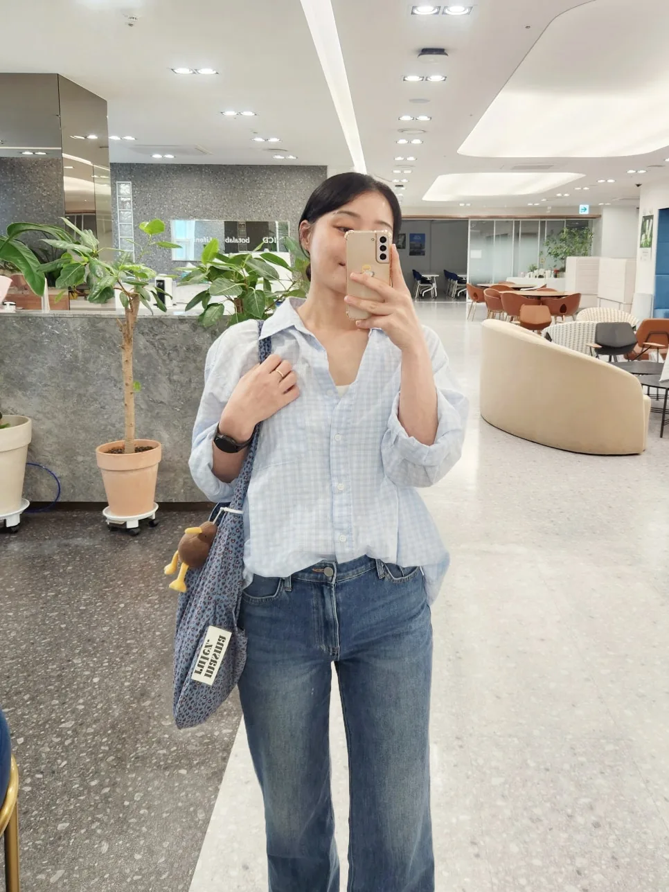 Woman taking a mirror selfie in a bright, air-conditioned office lobby with plants and lounge seating